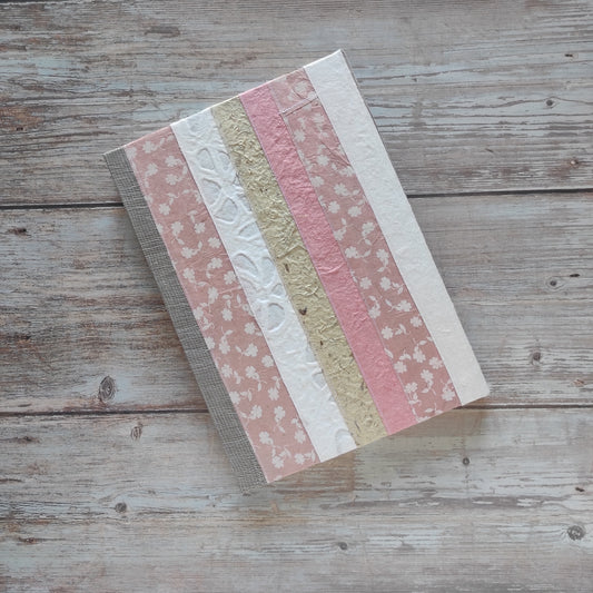 Diario rilegato con copertina rigida rivestita con un patchwork di carte artigianali cucite tra loro. Tonalità rosa, bianco e naturale.