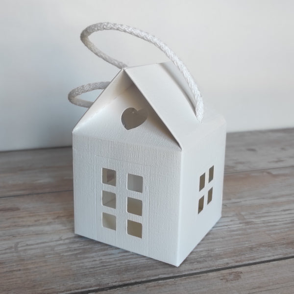 Scatola-Lanterna a forma di casetta in cartoncino fibra colore bianco. Misura 8 cm x 8 cm, H 12 cm ed è completa di manico in corda.