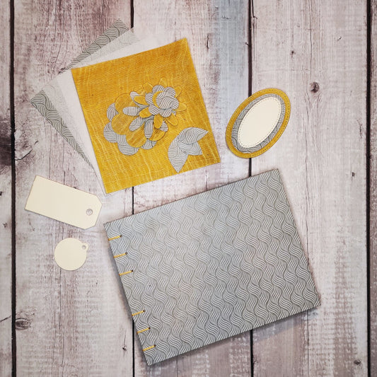 Album portafotografie realizzato a mano con la tecnica della rilegatura copta. La copertina è rivestita con carta Lokta (produzione artigianale nepalese) stampata con un motivo ad onde chiaro su fondo grigio-azzurro (River Bluegrey/White) e cucitura a vista in giallo. L'album ha 20 fogli (40 facciate) in cartoncino avorio 230 gr/m² e 1 tasca con soffietto in cartoncino giallo contenente degli elementi in carta che possono essere utilizzati per decorare le pagine: ritagli, tags, etichette, fiori e foglie.