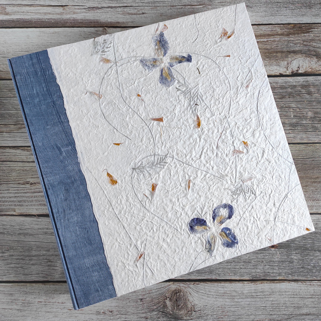Copertina album fotografico grande formato in carta di gelso con fiori blu e petali gialli grandi inserti su fondo avorio.